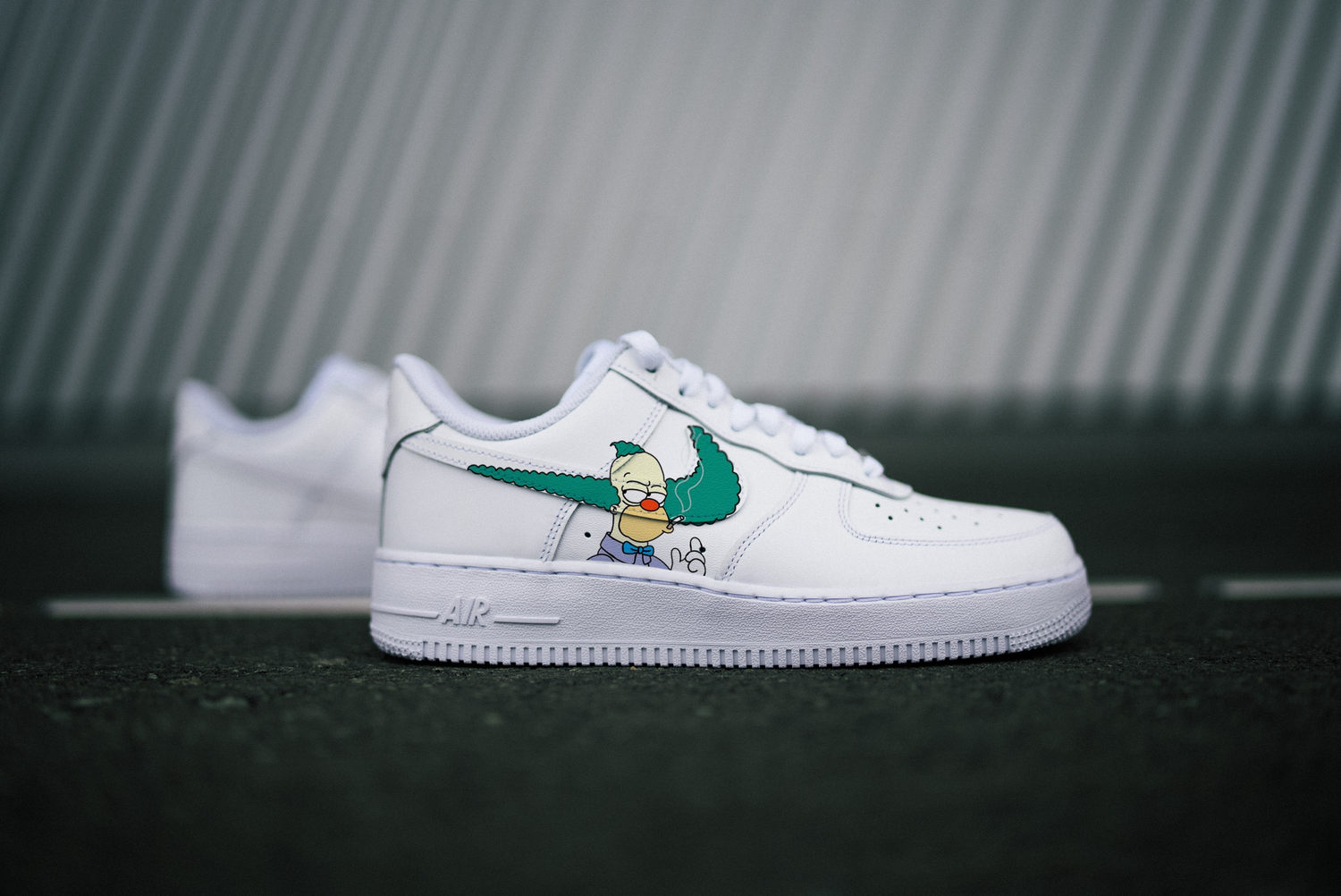The image is featuring a Custom Nike Air force 1 sneakers on a gray asphalt background. On the side of the sneaker is a design of krusty the clown from simpsons.