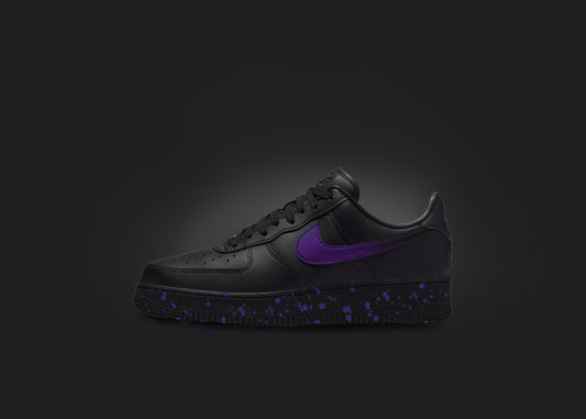 The image is featuring a custom black Air force 1 sneaker on a blank black background. The black nike sneaker has a purple paint splatter design on the sole and a purple fade on the nike logo. 