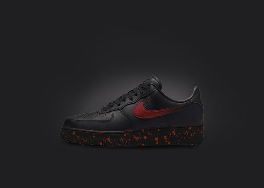 The image is featuring a custom black Air force 1 sneaker on a blank black background. The black nike sneaker has a red paint splatter design on the sole and a red fade on the nike logo. 