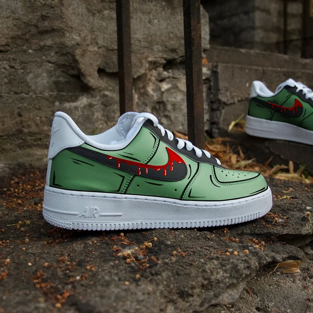 The image is featuring a Custom green cartoon Air force 1 sneaker pair on a street background. The white nike sneakers have a green cartoon design on the entire air force sneakers. 