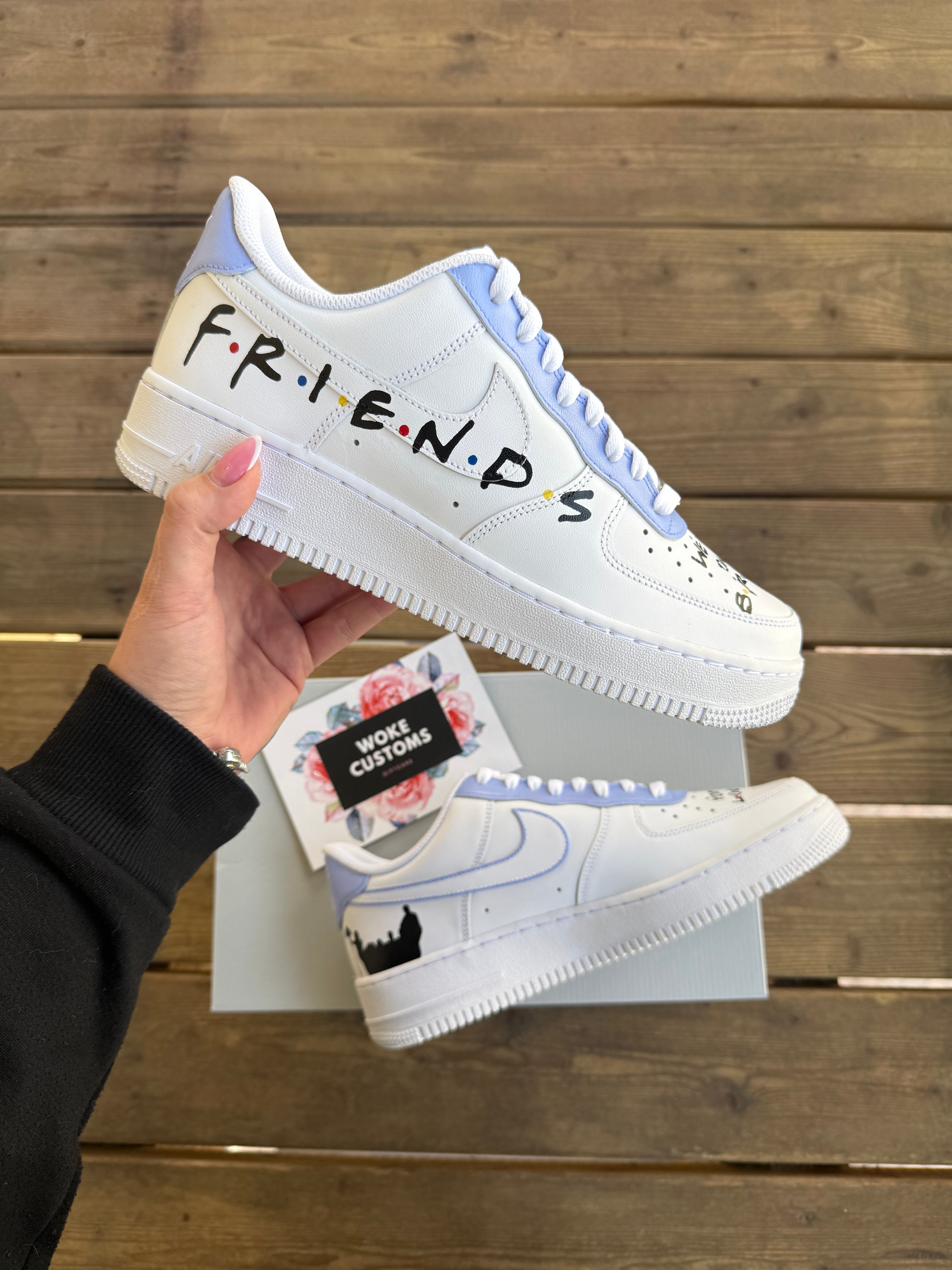 white nike air force 1 sneakers, with a friends tv series themed logo design covering the side of the sneaker. brown wooden background, a hand holding a single sneaker in the air