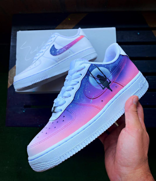 A custom Nike Air Force 1 sneaker featuring a pink and purple galaxy-themed design with a silhouette of a person on a swing in front of a large moon, adding a dreamy effect. The background blends into shades of space, speckled with white stars, creating a nighttime aesthetic.
