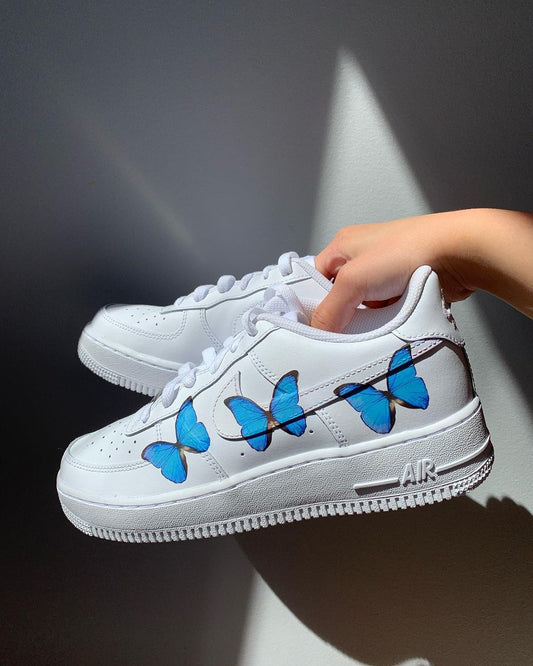 a pair of nike air force 1s with a simple blue butterfly design on the outside sections of the sneakers. Blank gray shadow wall on the background