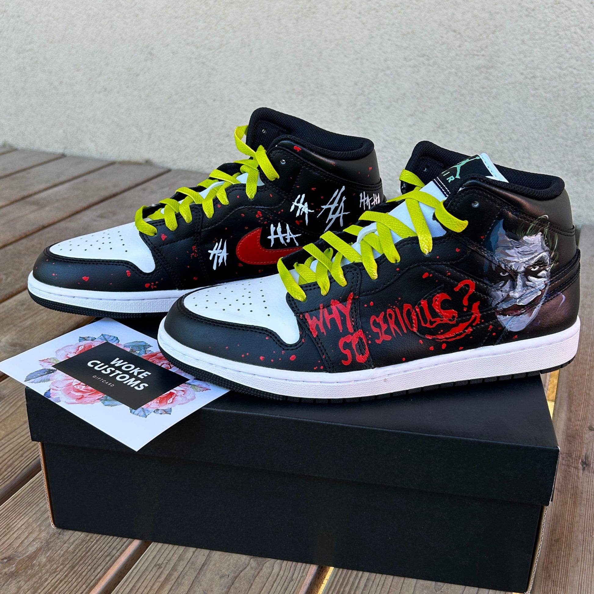 Custom hand painted air jordan 1 mid sneakers with a joker themed design. Sneakers are moslty in black with a white sole placed on a black sneakerbox, from a side view. The design have ajoker portrait on the closer sneaker with text "why so serious". On the right sneaker behind the left sneaker the design has joker themed Ha Ha Ha's on it, written in white. Both sneakers have red nike logos and red paint splatters on the sides. 