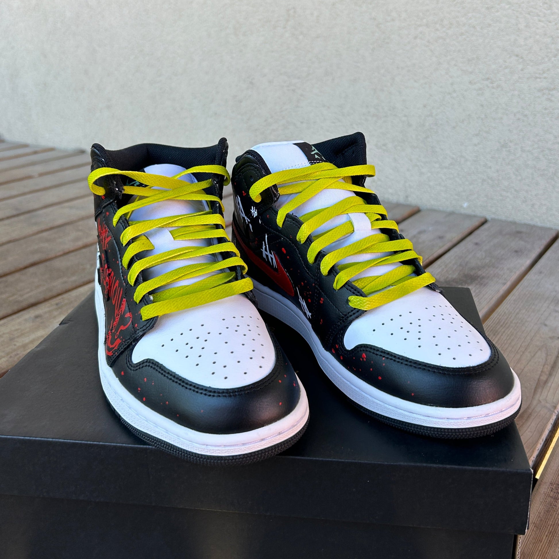 two jordan 1 sneakers placed on a black shoebox, picture taken from the toe sides. Sneakers are black and white with subtle red splatter on the black sections. Sneakers also have lime colored laces. 