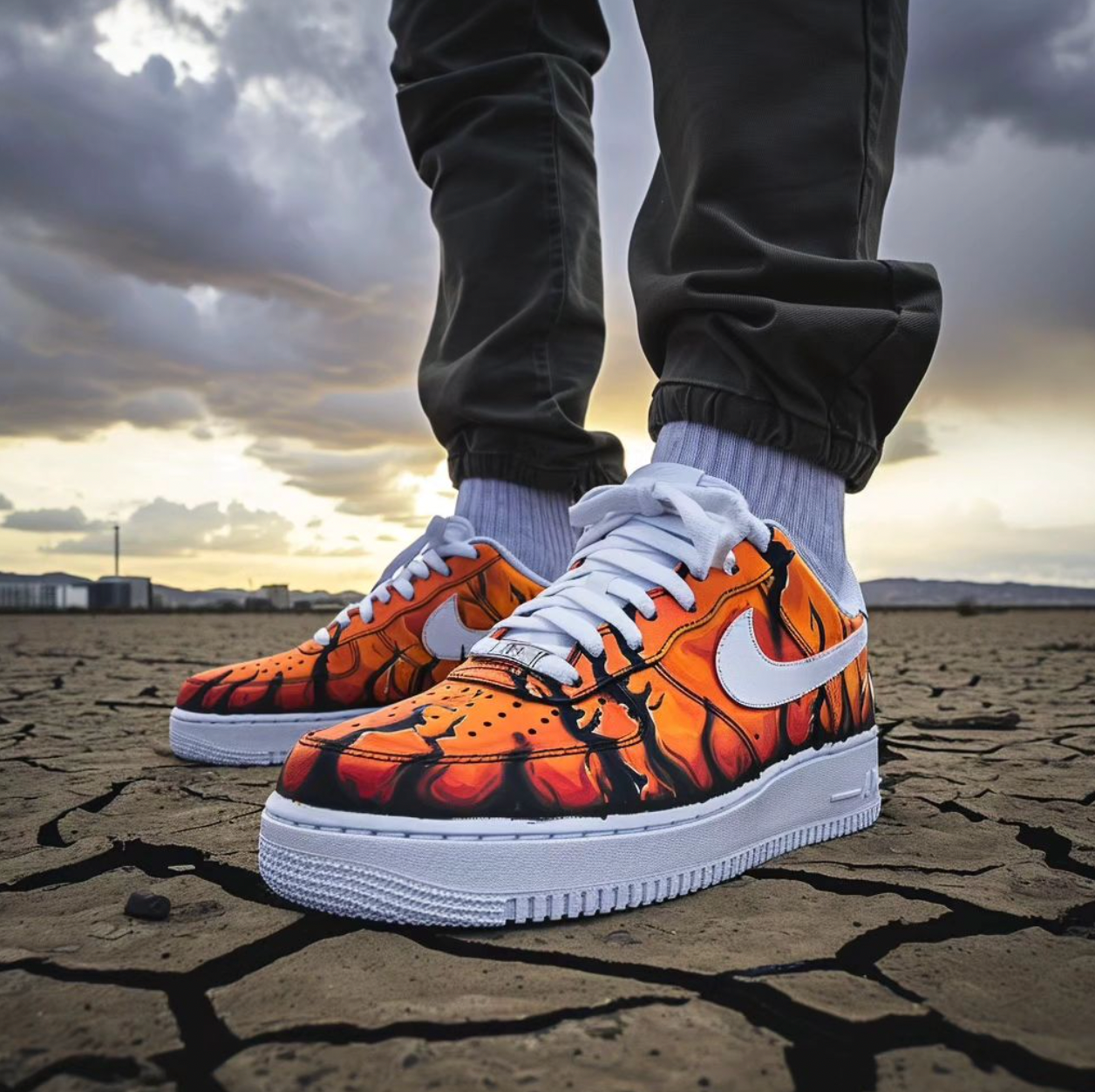 A person wearing customized Nike sneakers with an orange lava design, standing on cracked, dry ground with a dramatic, cloudy sky in the background.