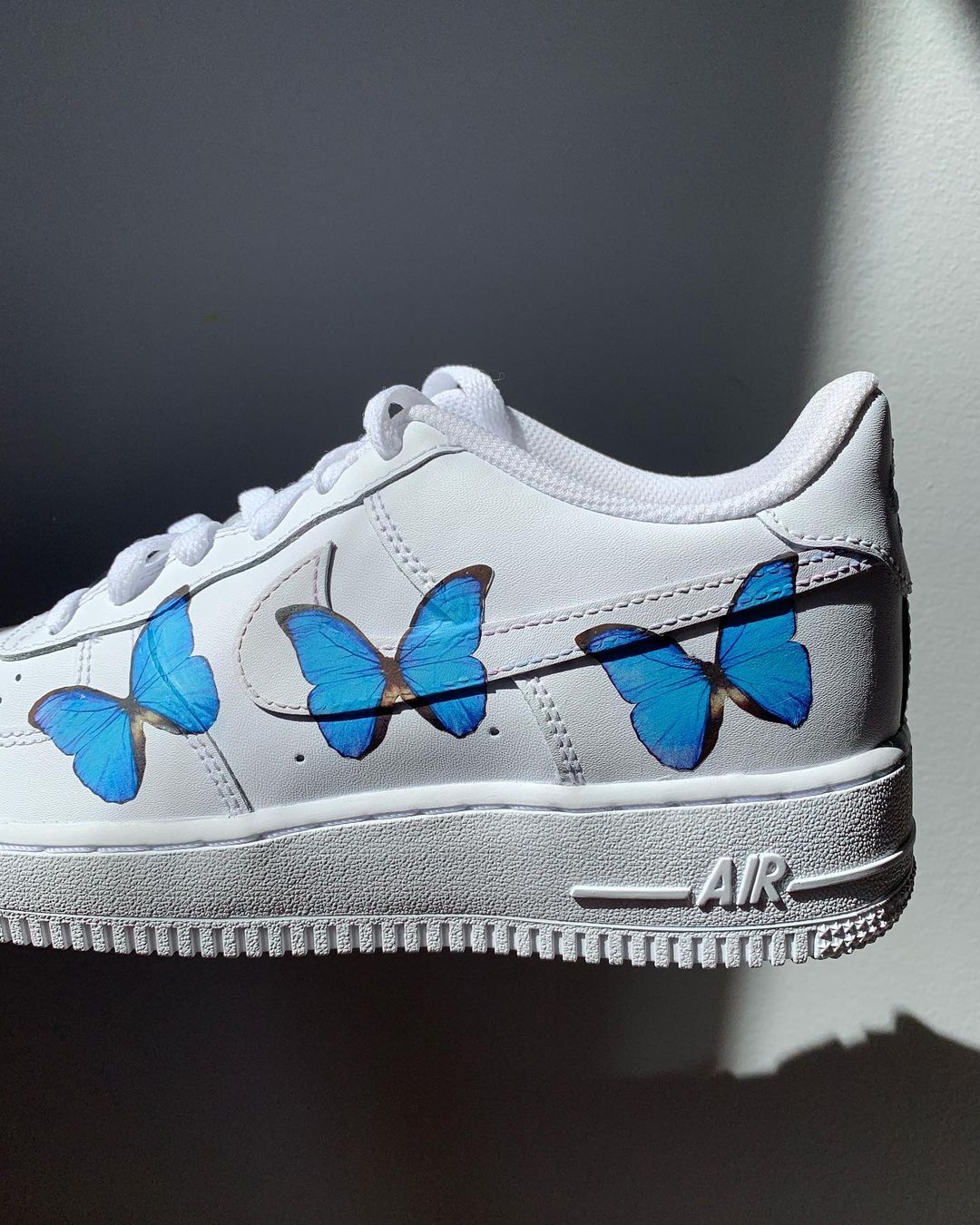 A single nike air force 1 sneakers with 3  blue butterfly designs on the outside sections of the sneakers. Blank gray shadow wall on the background