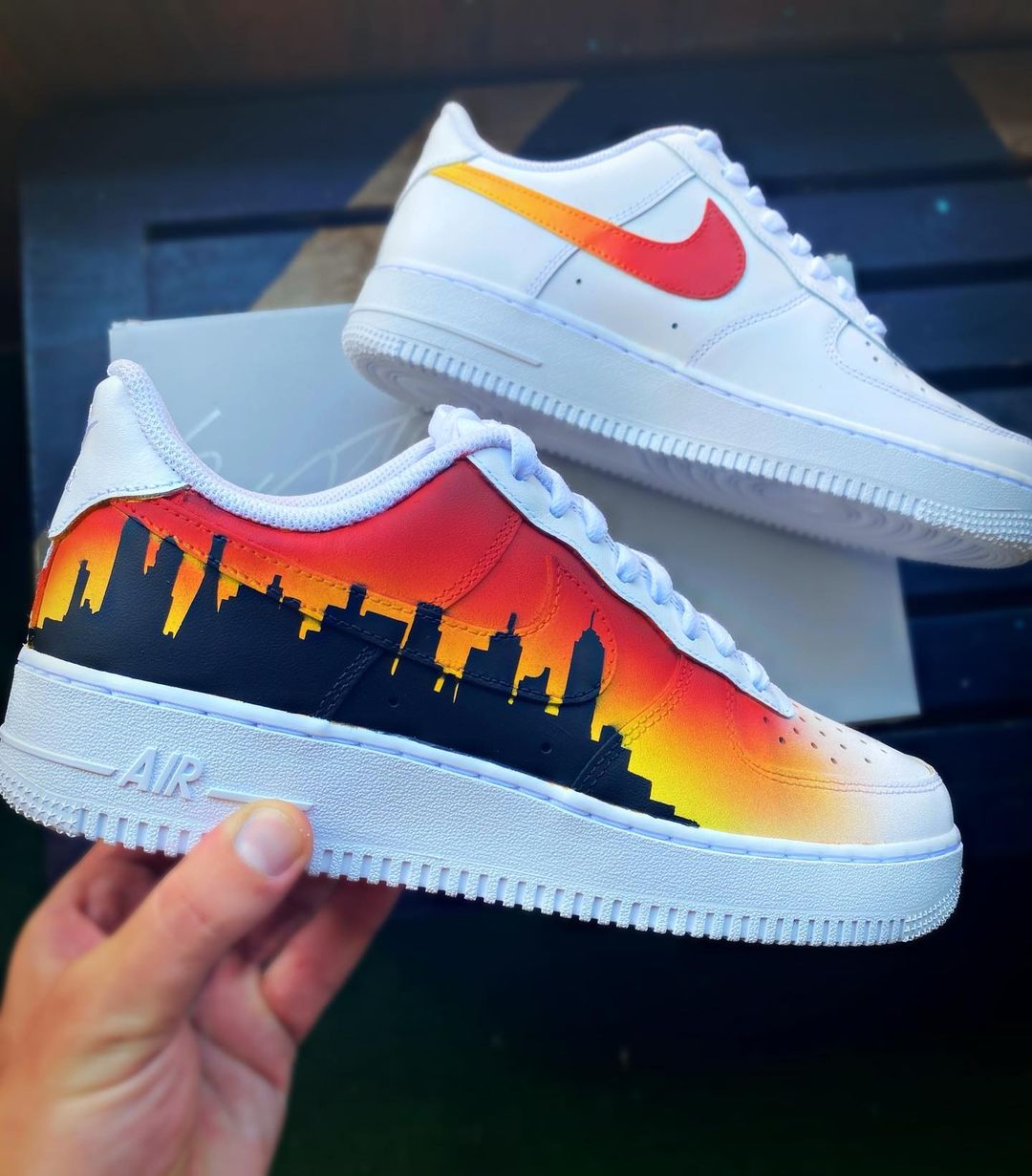 Nike Air Force 1 with a custom painted cityscape silhouette against a vibrant orange and yellow sunset gradient.