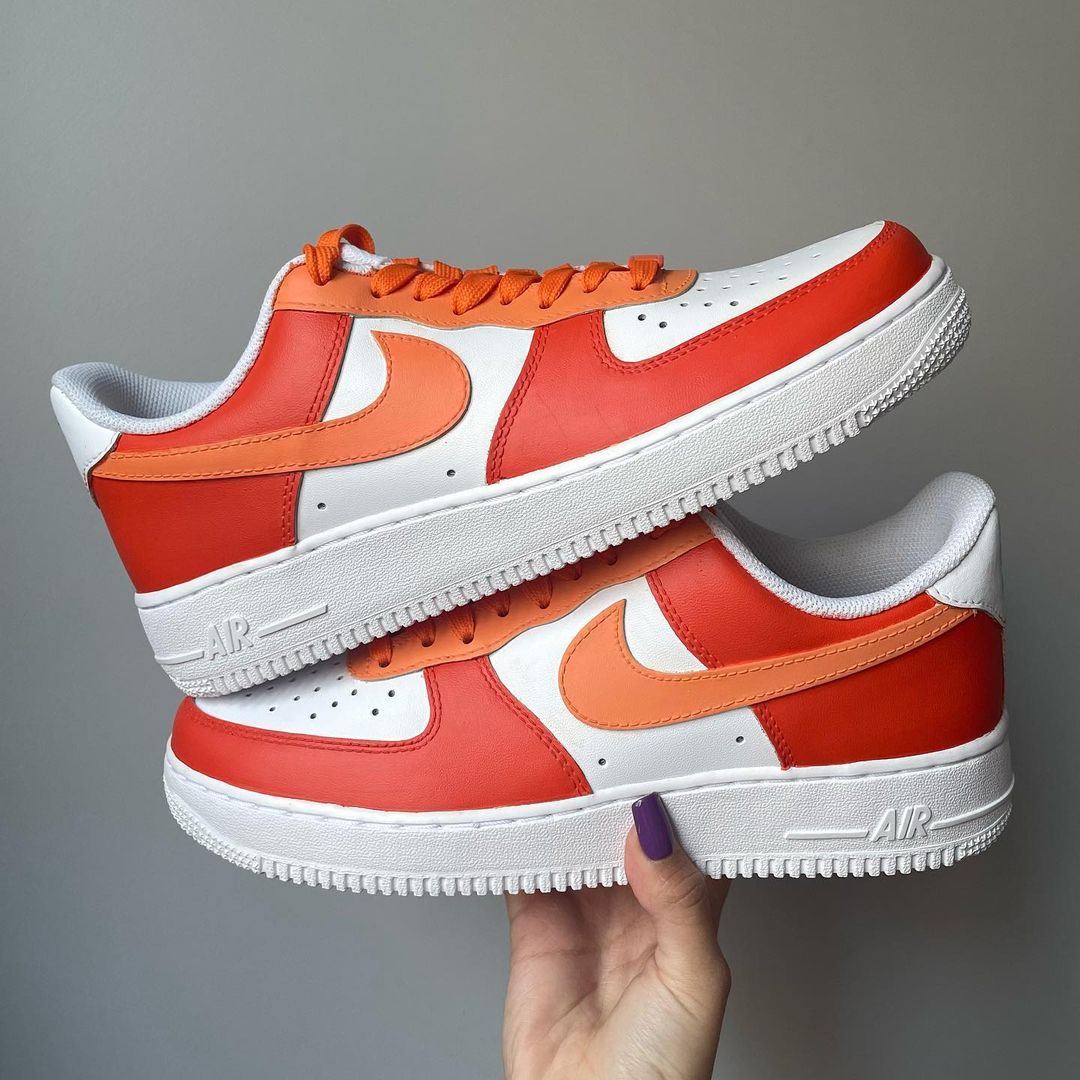 a pair of nike air force 1s with a orange tones design on the outside sections of the sneakers. Orange laces. Blank gray shadow wall on the background
