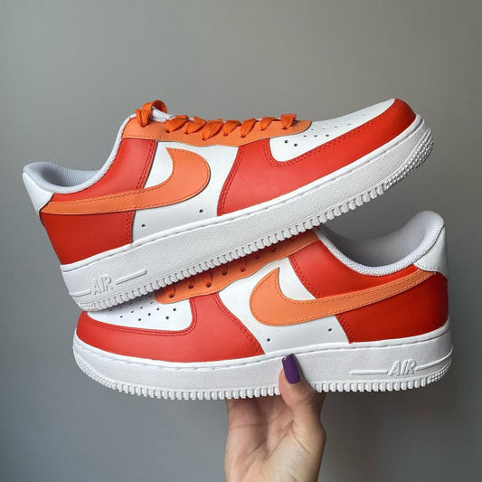 a pair of nike air force 1s with a orange tones design on the outside sections of the sneakers. Orange laces. Blank gray shadow wall on the background