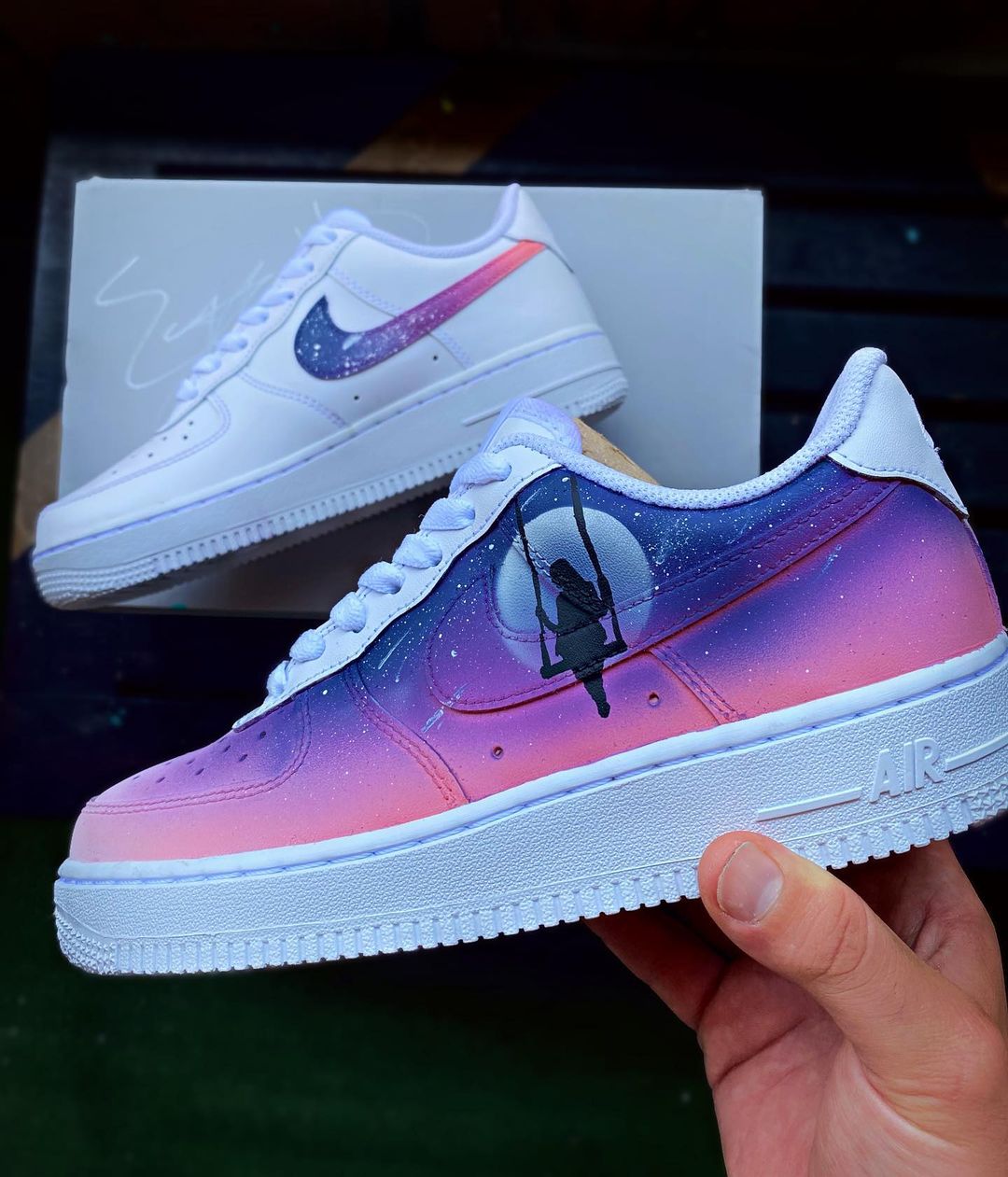 Side view of a custom-painted Nike Air Force 1 shoe with a serene night sky design, showcasing a figure on a swing in front of a full moon. The sneaker features soft pink and purple gradients with starry accents to create a cosmic vibe.