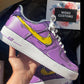 A custom-designed Nike Air Force 1 sneaker is displayed, being held in hand. The shoe features a vibrant purple color with a bold, yellow Nike swoosh, outlined with black lines creating a comic book style. In the background, a Woke Customs gift card, adorned with a floral design, is placed on black wrapping paper.