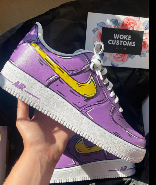 A custom-designed Nike Air Force 1 sneaker is displayed, being held in hand. The shoe features a vibrant purple color with a bold, yellow Nike swoosh, outlined with black lines creating a comic book style. In the background, a Woke Customs gift card, adorned with a floral design, is placed on black wrapping paper.