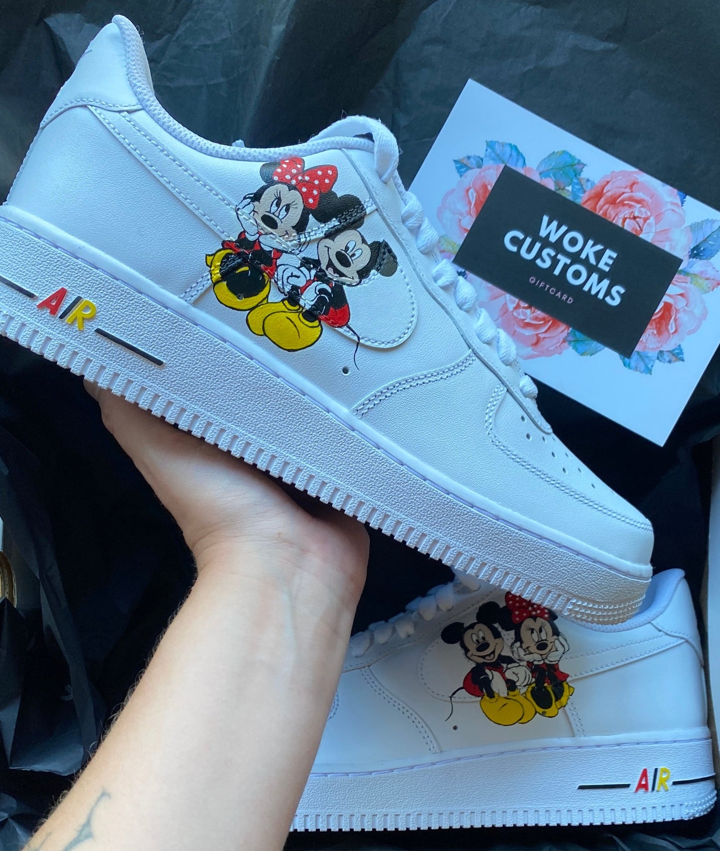 A pair of white custom Nike Air Force 1 sneakers featuring a hand-painted design of Mickey and Minnie Mouse on the side. The shoes have colorful accents on the “AIR” text near the sole. A hand is holding one of the sneakers, and a “WOKE CUSTOMS” gift card is visible in the background.