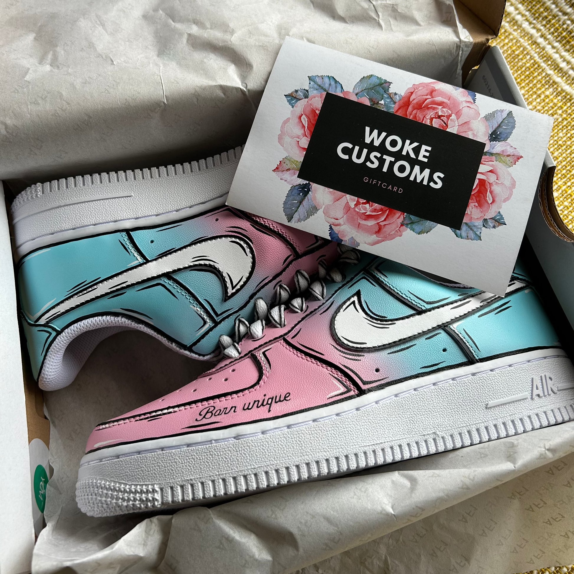 A pair of custom-designed Nike Air Force 1 sneakers are displayed in a shoebox. The shoes feature a unique pink and blue fade color scheme, with both colors blending seamlessly together, outlined with bold, black lines creating a comic book style. The phrase “Born unique” is inscribed on the side of the pink shoe. A Woke Customs gift card, adorned with a floral design, is placed on top of the shoes.