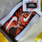 A pair of custom-designed Nike Air Force 1 sneakers are displayed in a shoebox. The shoes feature a hand-painted orange and black lava design, with a crisp white Nike swoosh. The sneakers are placed on white tissue paper, and a Woke Customs gift card with a floral design is positioned next to them. The yellow background adds a vibrant touch to the overall presentation.