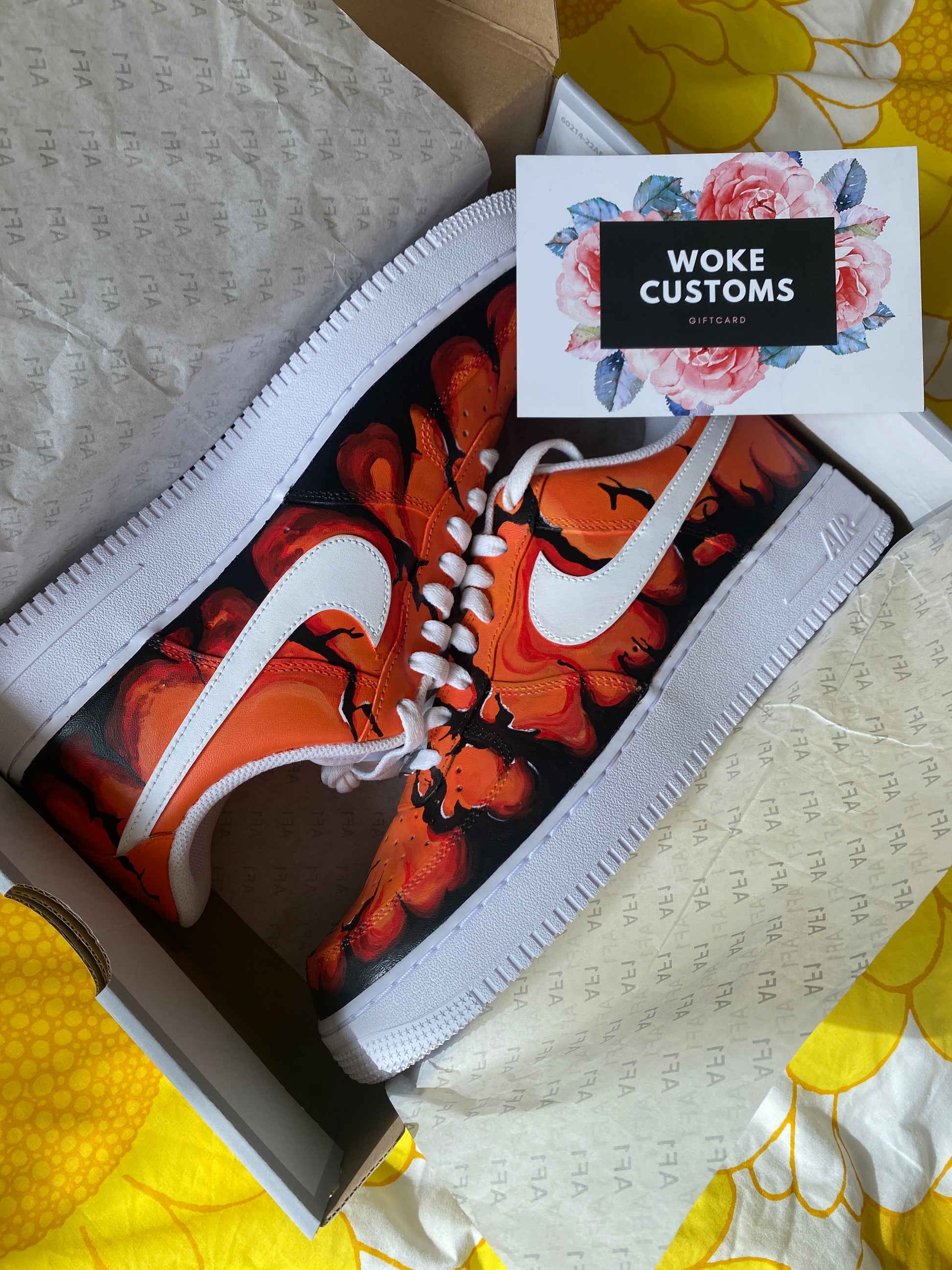 A pair of custom-designed Nike Air Force 1 sneakers are displayed in a shoebox. The shoes feature a hand-painted orange and black lava design, with a crisp white Nike swoosh. The sneakers are placed on white tissue paper, and a Woke Customs gift card with a floral design is positioned next to them. The yellow background adds a vibrant touch to the overall presentation.