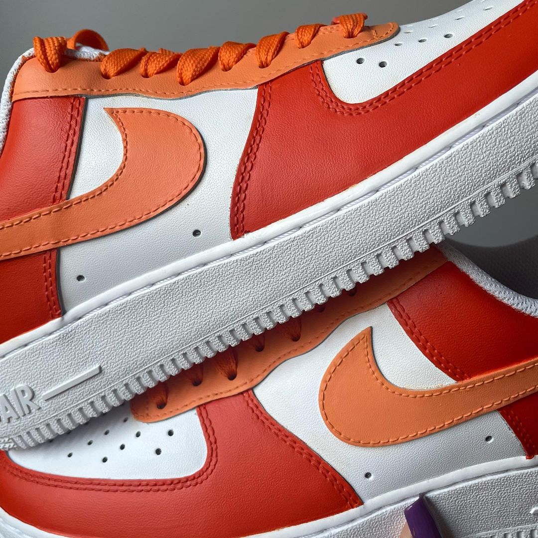 a closeup of a pair of nike air force 1s with a orange tones design on the outside sections of the sneakers. Orange laces. Blank gray shadow wall on the background