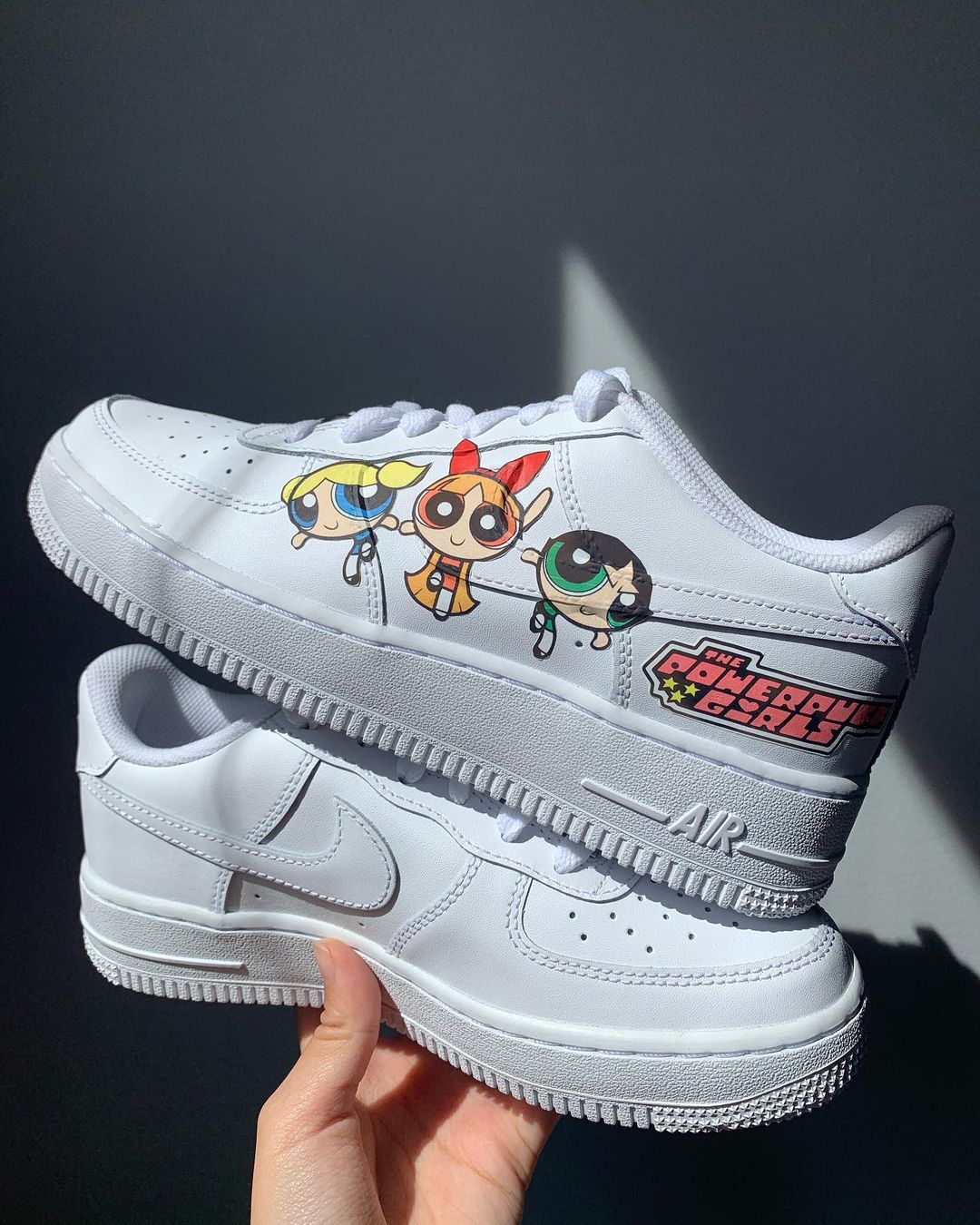 a pair of white nike air force 1s with a powerpuff girls design on the outside sections of the sneakers. Blank gray shadow wall on the background