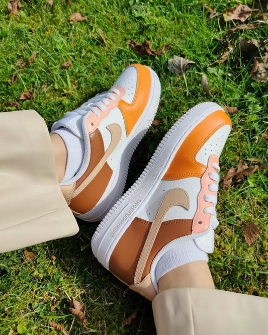 A pair of custom Nike Air Force 1 sneakers in warm autumn tones, featuring a combination of burnt orange and beige panels, captured on green grass. The hand-painted design accentuates a cozy fall aesthetic.