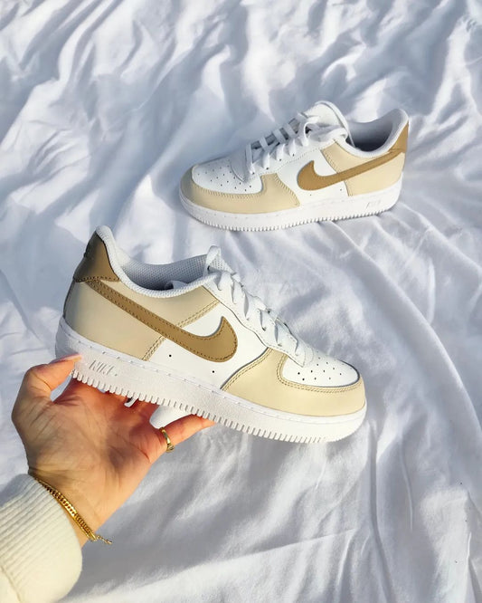 A pair of custom Nike Air Force 1 sneakers in soft beige and white tones, displayed on a white fabric background. The sneakers feature hand-painted beige swooshes and overlay panels, creating a minimalist, neutral aesthetic that complements the clean lines of the classic Air Force 1 design.