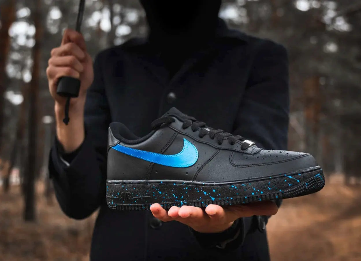 a black nike air force 1 sneakers, placed on a hand in the nature. The sneakers have a light blue paint splatter across the soles of the sneaker and a simple ombre fade design on the side nike logo swooshes. 