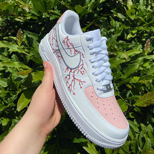 Hand-painted Air Force 1 sneakers featuring a delicate cherry blossom design in pink and white, accented with a soft pink toe cap.
