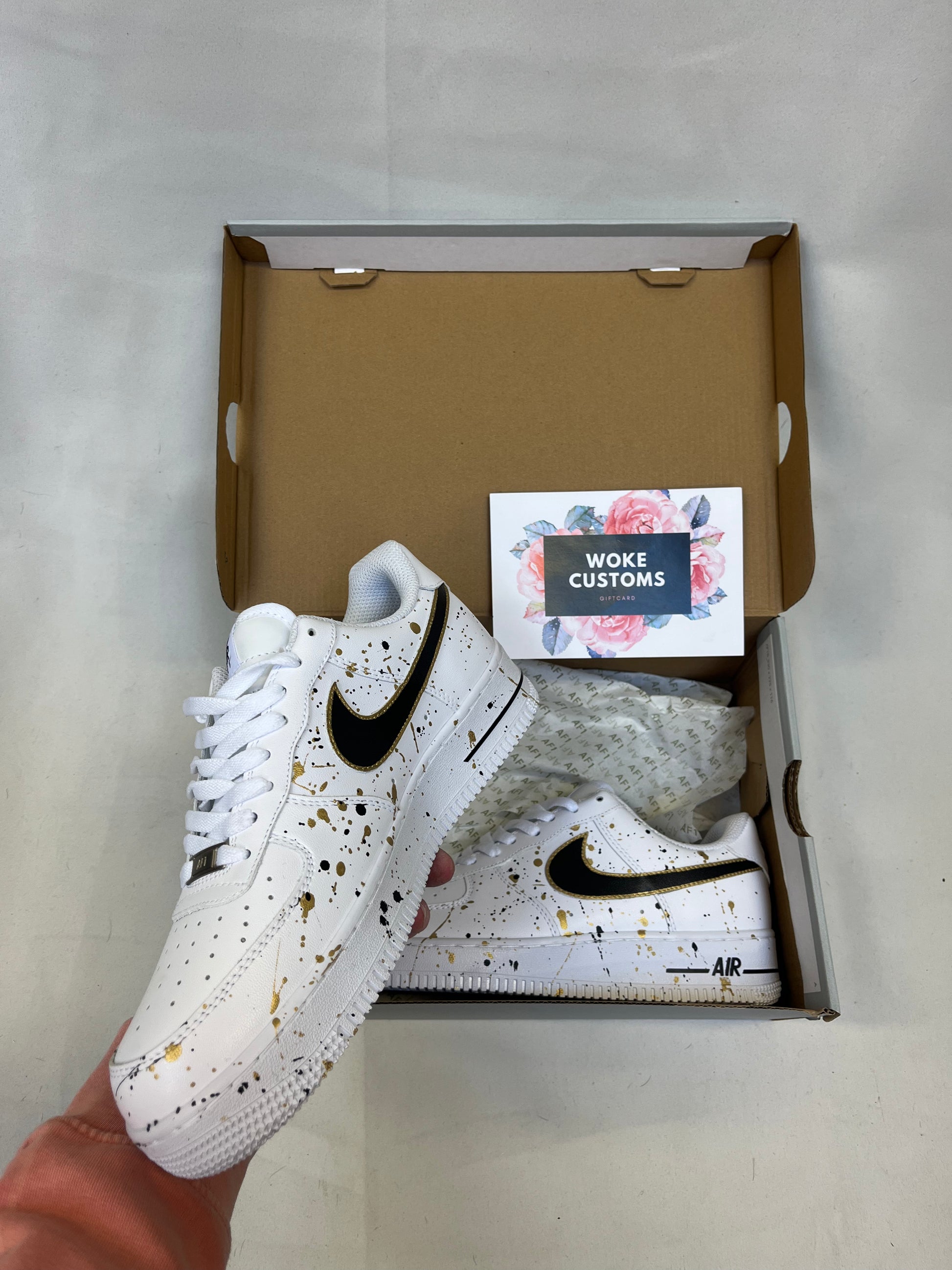 A pair of custom white Air Force 1 sneakers with elegant black and gold splatter design, handpainted custom white Nike shoes.