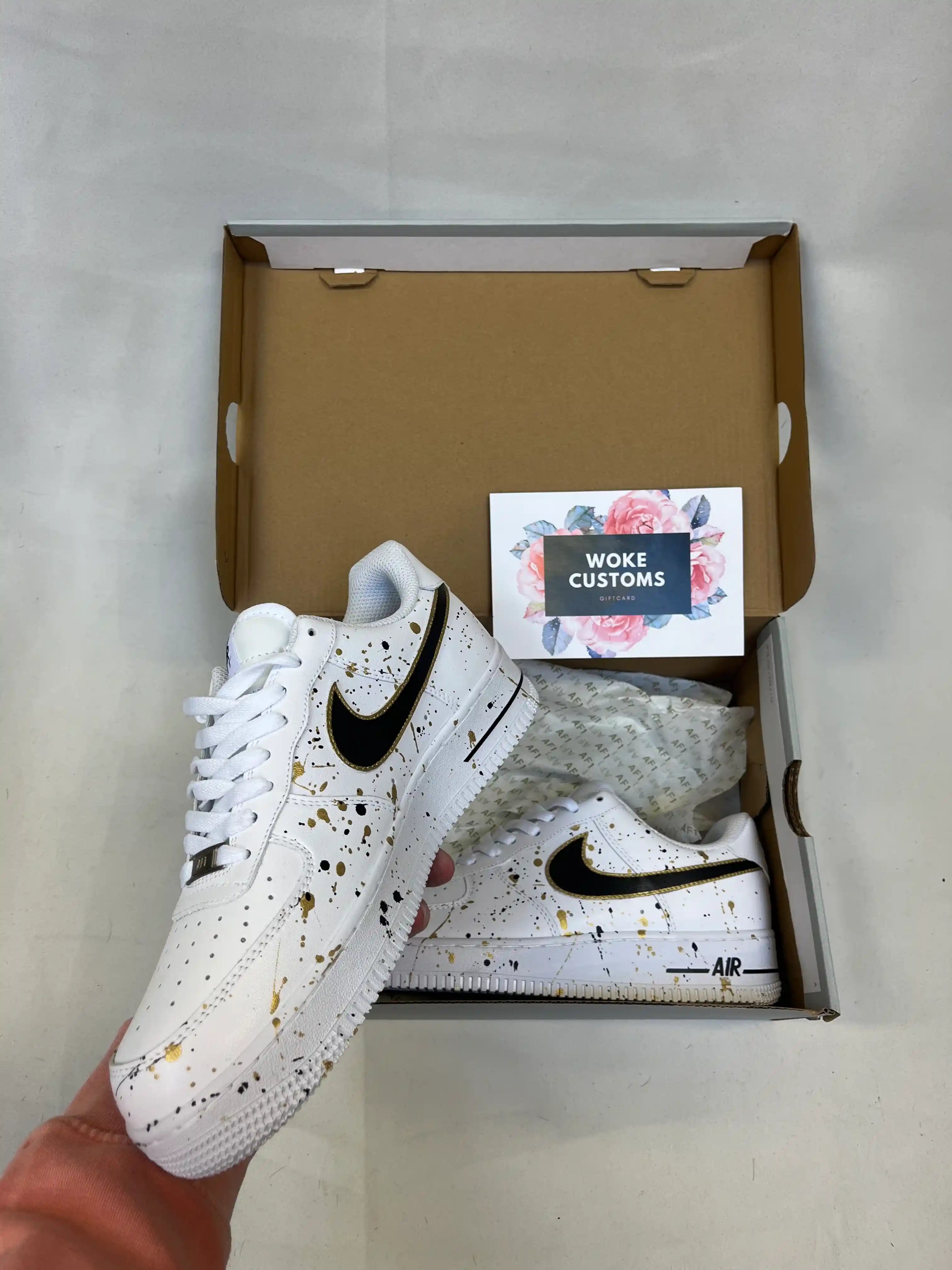A pair of custom white Air Force 1 sneakers with elegant black and gold splatter design, handpainted custom white Nike shoes.