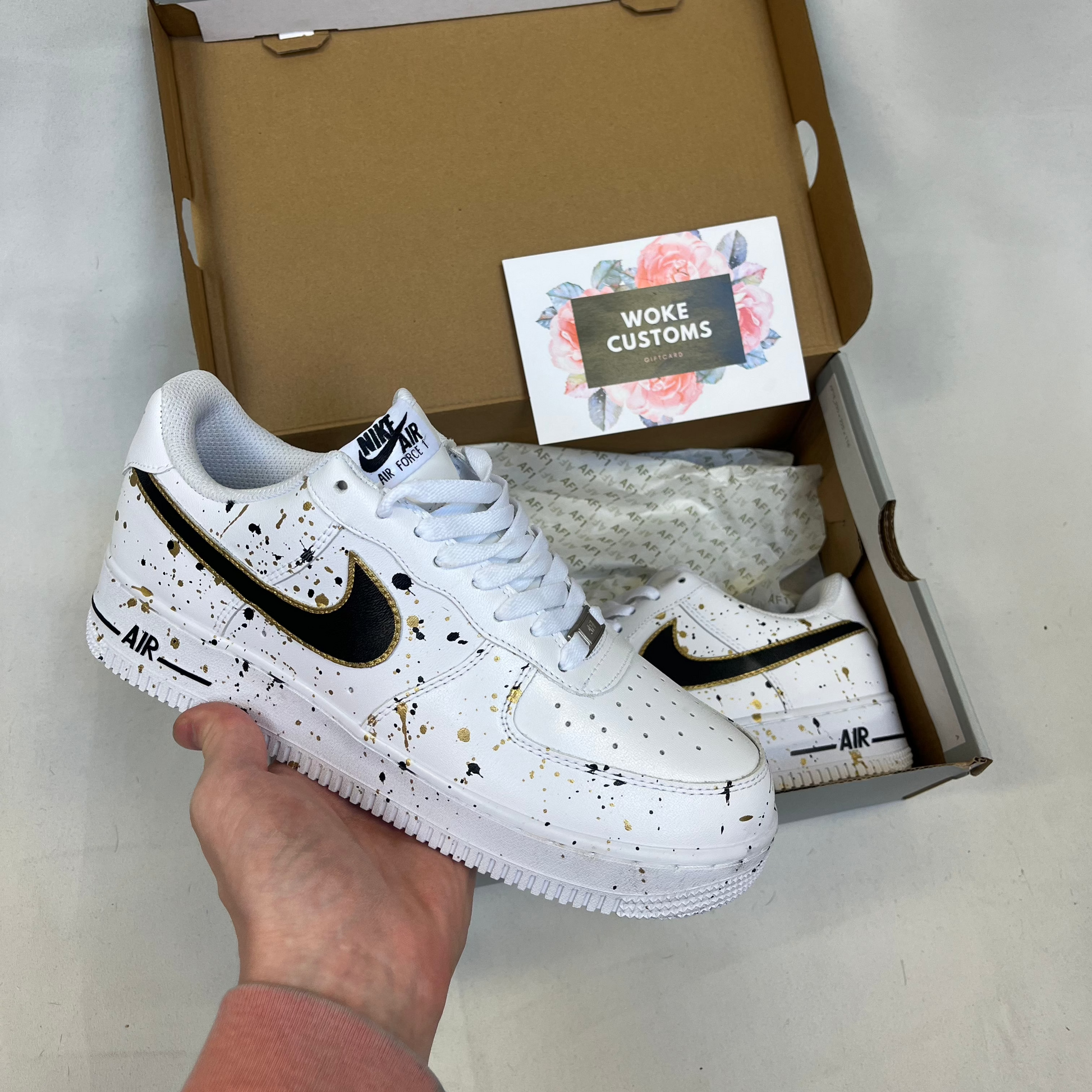 White custom Air Force 1s featuring artistic black and gold paint splatter pattern, premium handpainted sneakers displayed in box with side swoosh detail