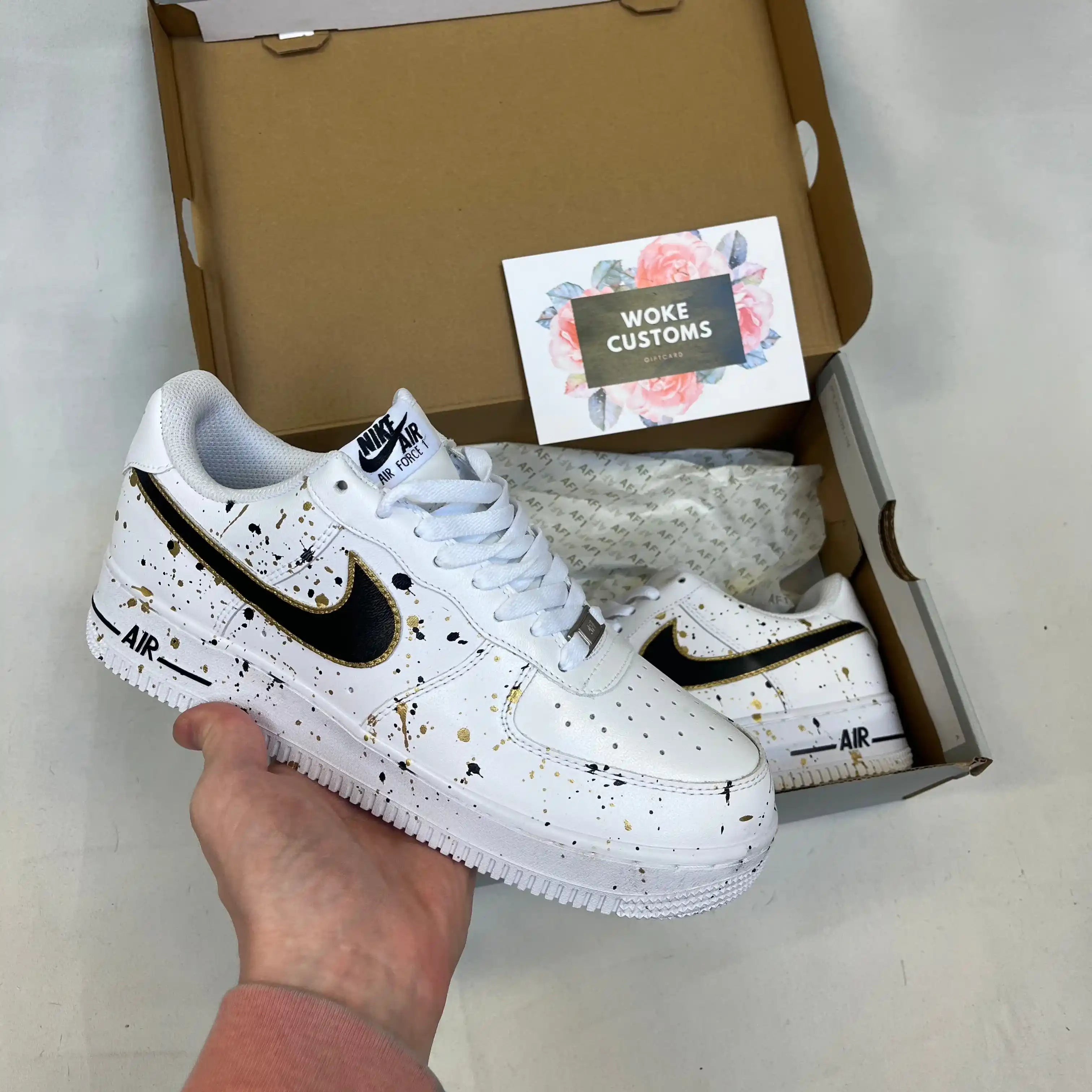 White custom Air Force 1s featuring artistic black and gold paint splatter pattern, premium handpainted sneakers displayed in box with side swoosh detail