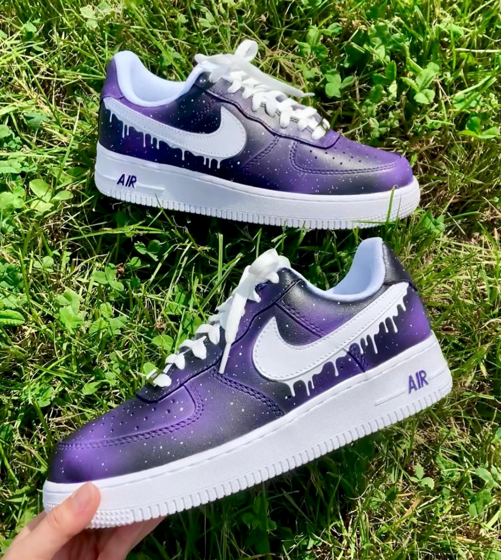 a pair of purple galaxy designed nike air force 1 sneaker with a white sole. The galaxy design covers the entire sneakers and features a white nike side logo swoosh with small white dripping details. the sneakers are placed on a grass background