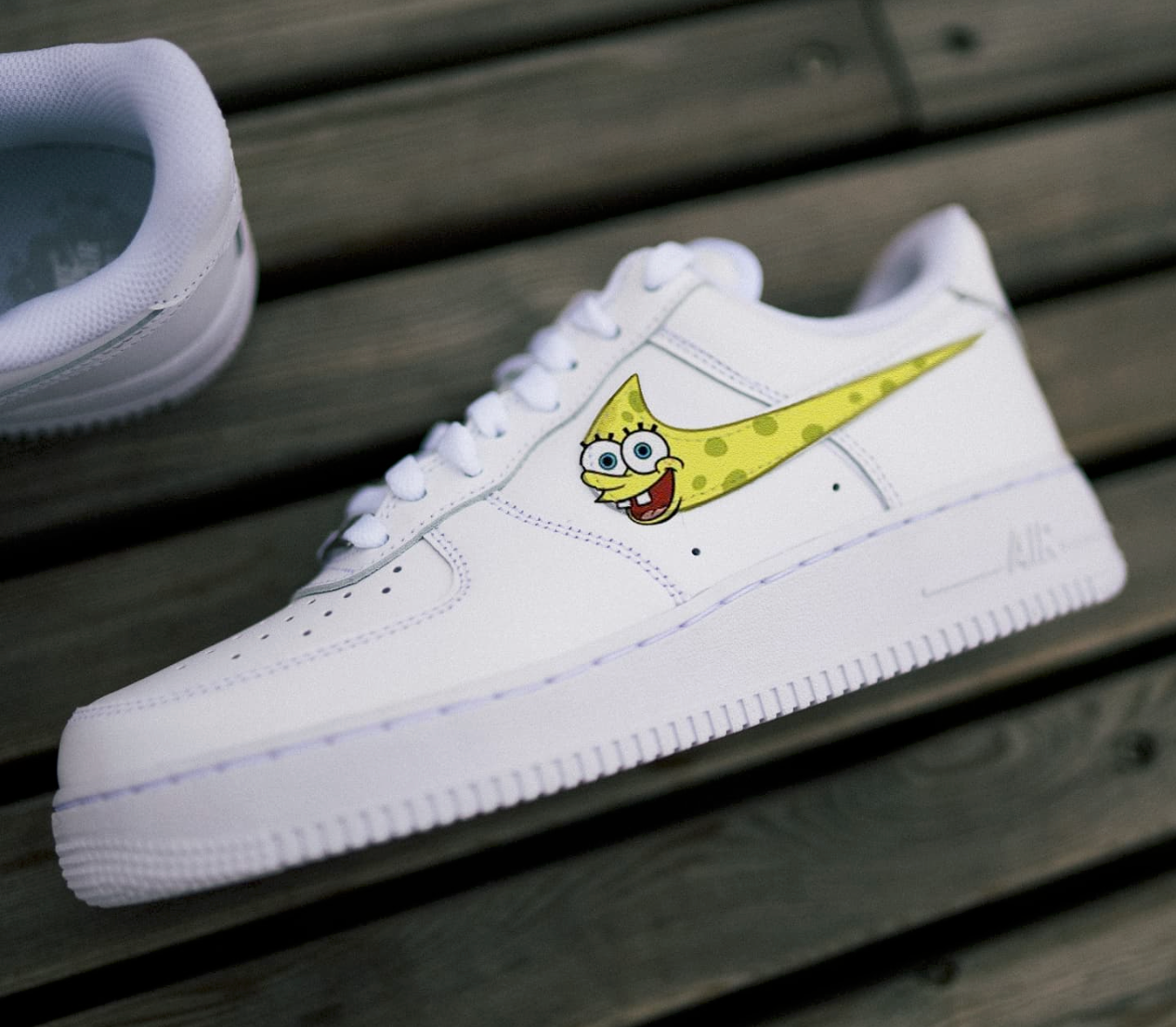 a single white nike air force 1 laying on its side on a park bench. the side nike logo has a spongebob design in yellow