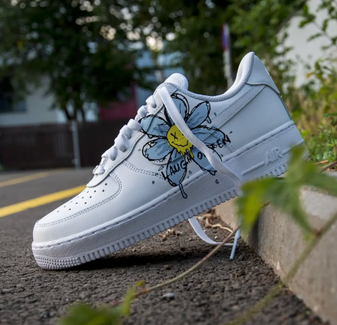 a hite nike air force 1 sneaker with a custom handpainted design of a blue flower and text laugh often on the side of the sneaker, the sneakers is placed in a street setting