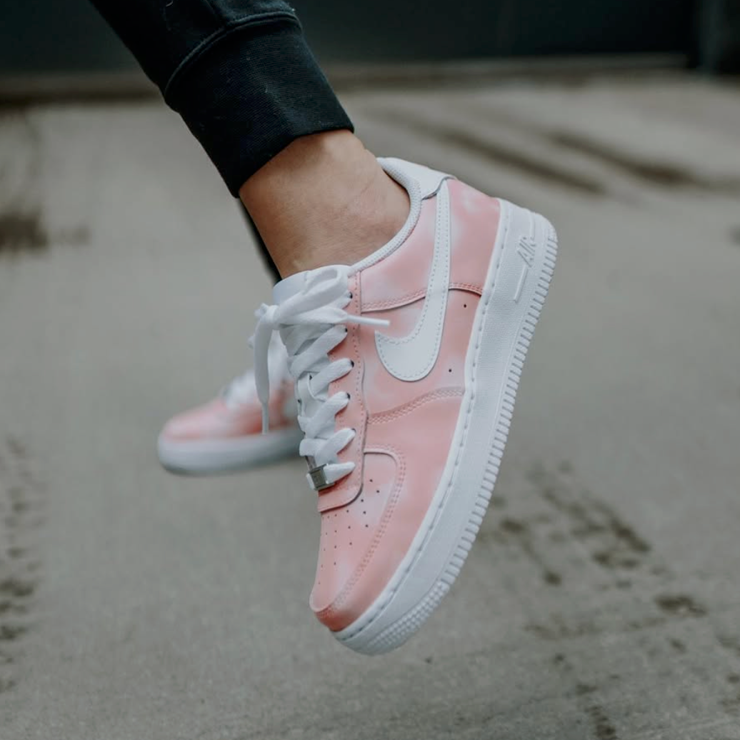 woman wearing and showing off a white nike air force 1 custom sneaker covered in a pink tone with lighter cloud like designs covering the sneaker. street setting