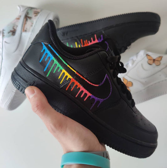 Black Nike Air Force 1 sneaker featuring a vibrant rainbow drip design on the Swoosh, with colors fading from blue to red. The sneaker is held up to showcase the colorful contrast against the matte black background, highlighting the unique hand-painted customization.