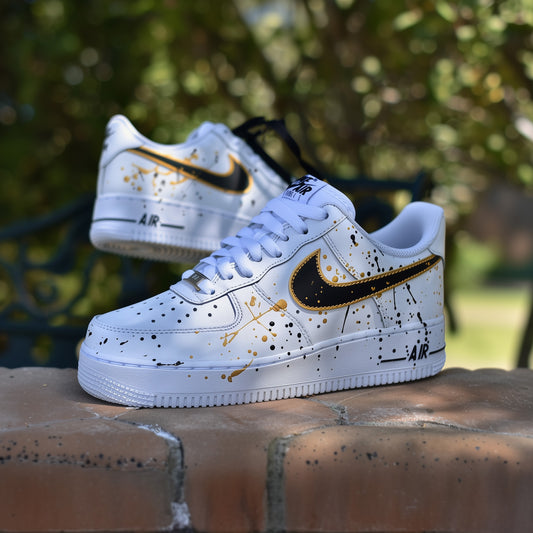 white nike air force 1s with a gold and black splatter desing on all sides of the sneakers. nike logo is painted in black with a gold outline. Paint splatter in gold and black covering the sides of the sneakers