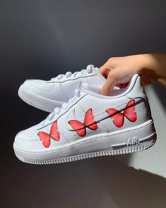 a pair of nike air force 1s with a simple red butterfly design on the outside sections of the sneakers. Blank gray shadow wall on the background
