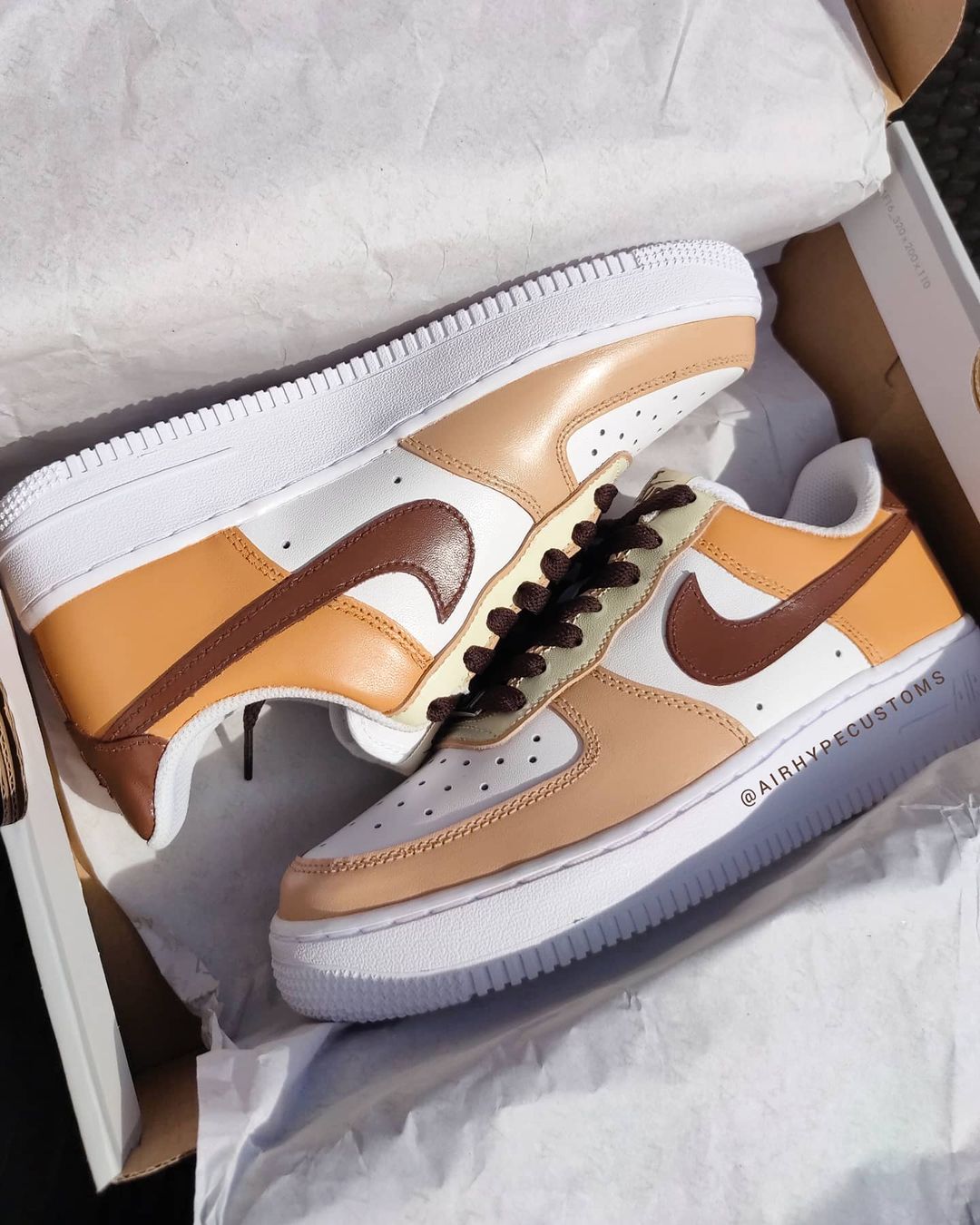 A pair of custom Nike Air Force 1 sneakers in a box, featuring a hand-painted design with earth tones of brown, beige, and tan on white leather, with brown laces adding a stylish contrast.