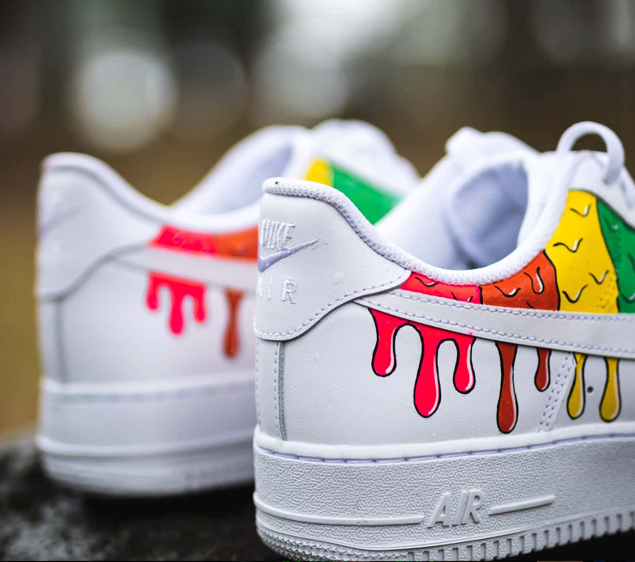 a pair of custom nike air force 1 white sneakers placed on a rock with colorful paint dripping design on the sides of the sneakers, closup heel view of the sneakers