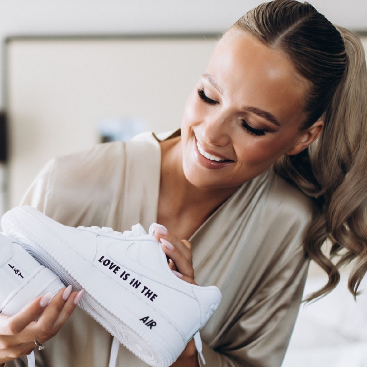 woman showing nike air force 1 white sneaker with text love is in the air in black written on the sneaker side