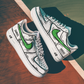 pair of white nike air force 1s with a cartoon design and a green nike logo. background is a tenniscourt