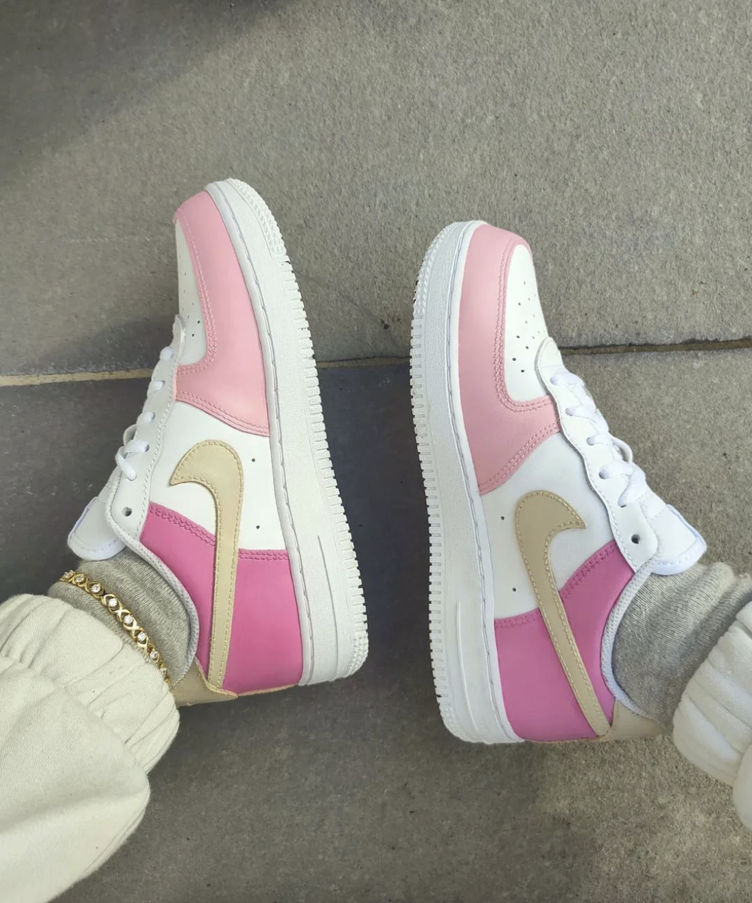 A pair of custom Nike Air Force 1 sneakers featuring soft pink and beige panels, creating a pastel aesthetic. The sneakers are styled on foot against a stone background, with the beige swoosh and pink overlays giving a unique, hand-painted look.