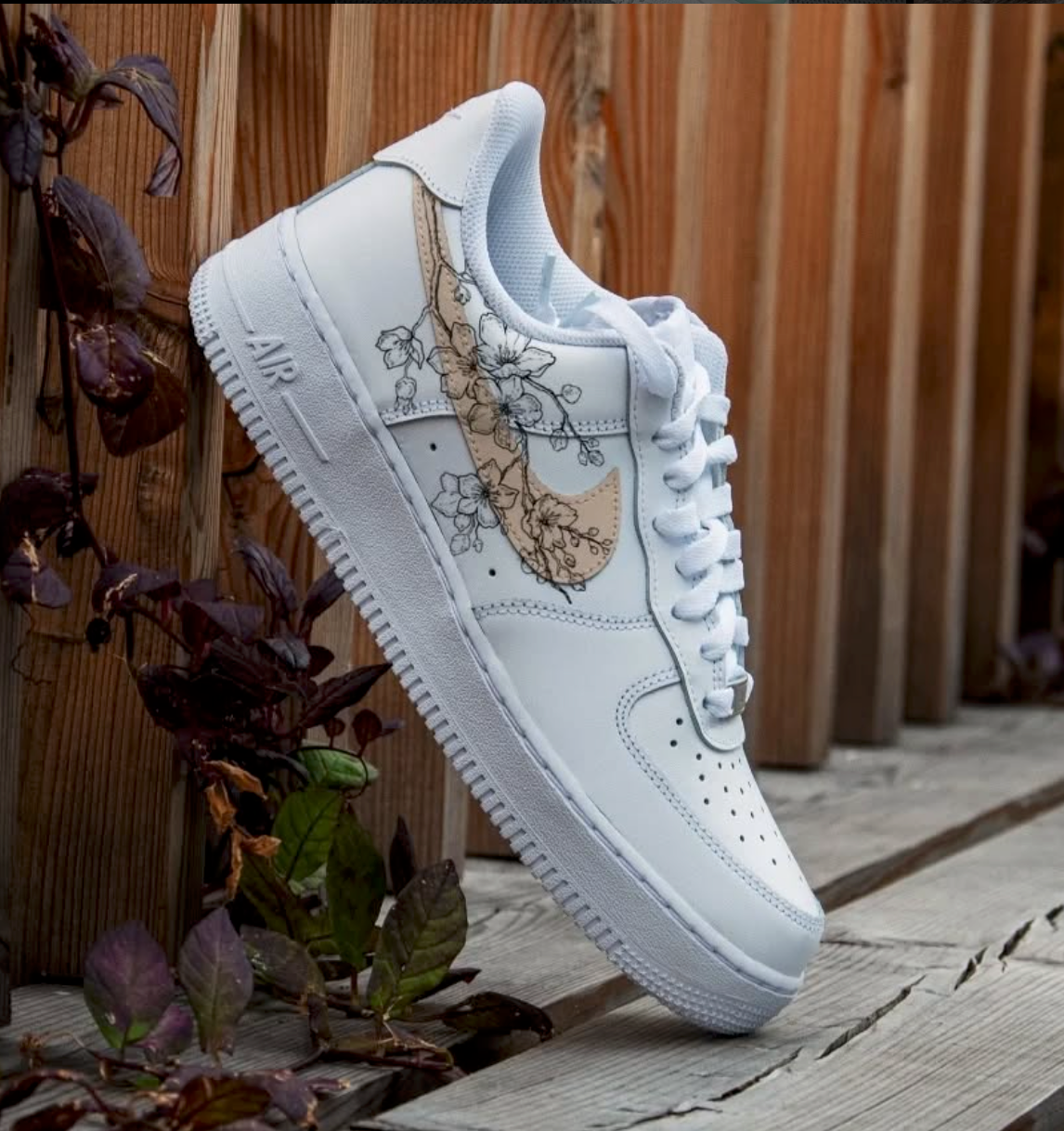 white nike air force 1 sneaker placed on a park bench side view. The sneaker feautres a minimal flower branch lineart covering a beige painted swoosh side nike logo. 