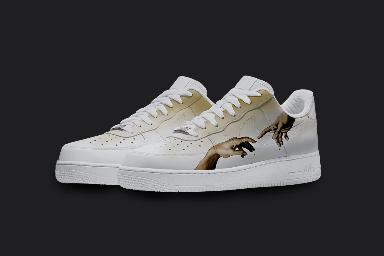 The image is of a Custom Nike Air force 1 sneaker pair on a blank black background. The white custom sneakers have a a brown fade design with a creation of adams hand illustrations on the sides of the sneaker. 