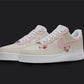 The image is of a Custom Nike Air force 1 sneaker pair on a blank black background. The white custom sneakers have a Pink nike logo with floral illustrations on the sides.