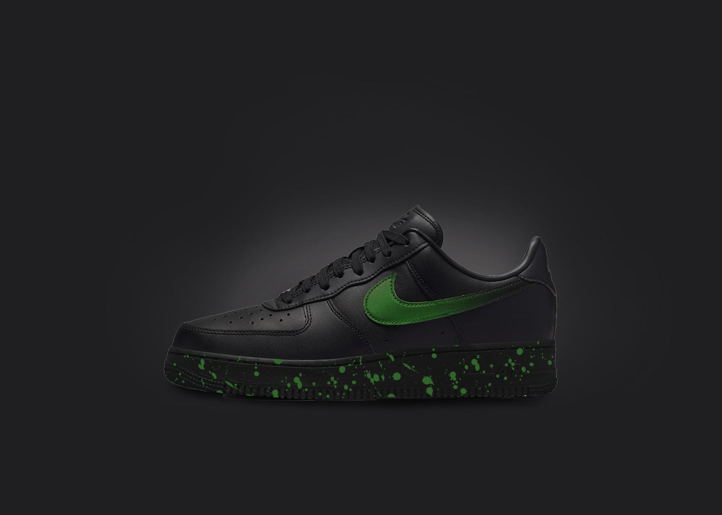 The image is featuring a custom black Air force 1 sneaker on a blank black background. The black nike sneaker has a green paint splatter design on the sole and a green fade on the nike logo. 