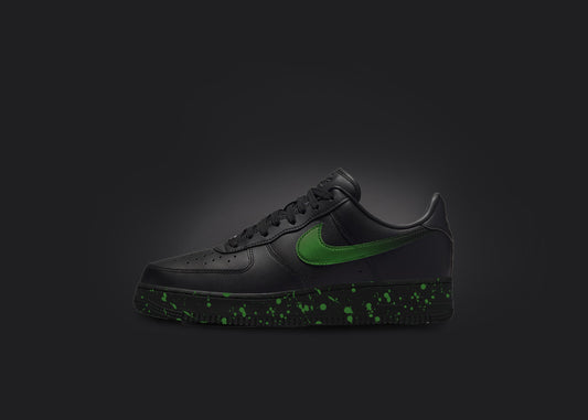 The image is featuring a custom black Air force 1 sneaker on a blank black background. The black nike sneaker has a green paint splatter design on the sole and a green fade on the nike logo. 